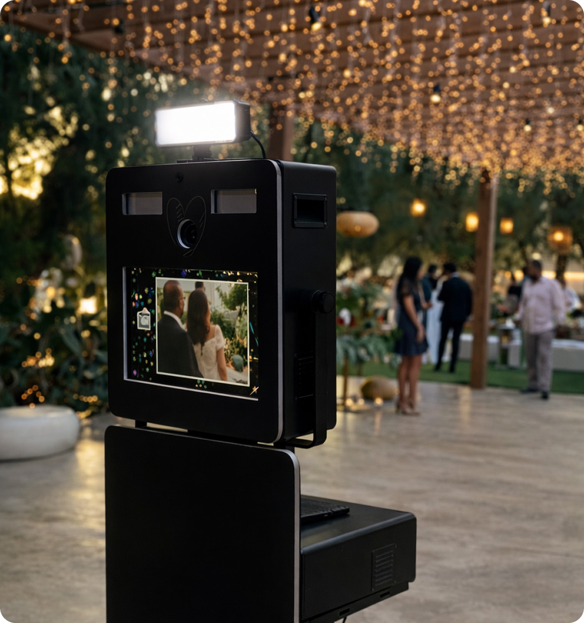 Photo booth capturing an image of a couple at an outdoor event with string lights overhead and blurred guests in the background.
