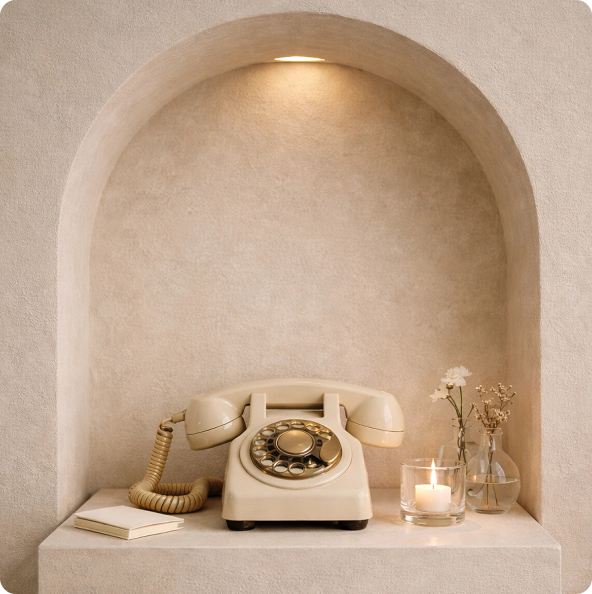 Beige rotary dial telephone placed on a shelf in a wall niche with a lit candle and small vases holding flowers.