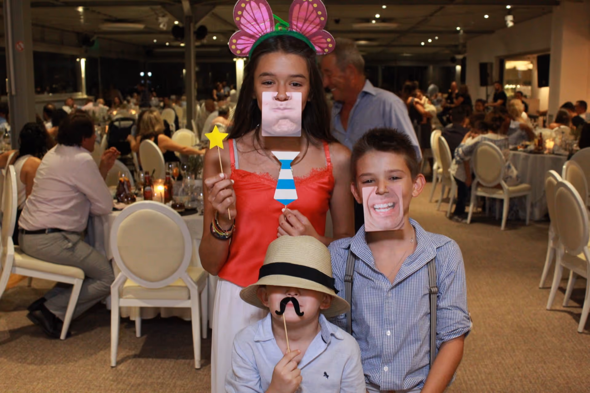 Three children posing indoors at a party with playful face and mustache masks, one girl wearing butterfly ears and holding a star wand and paper tie.