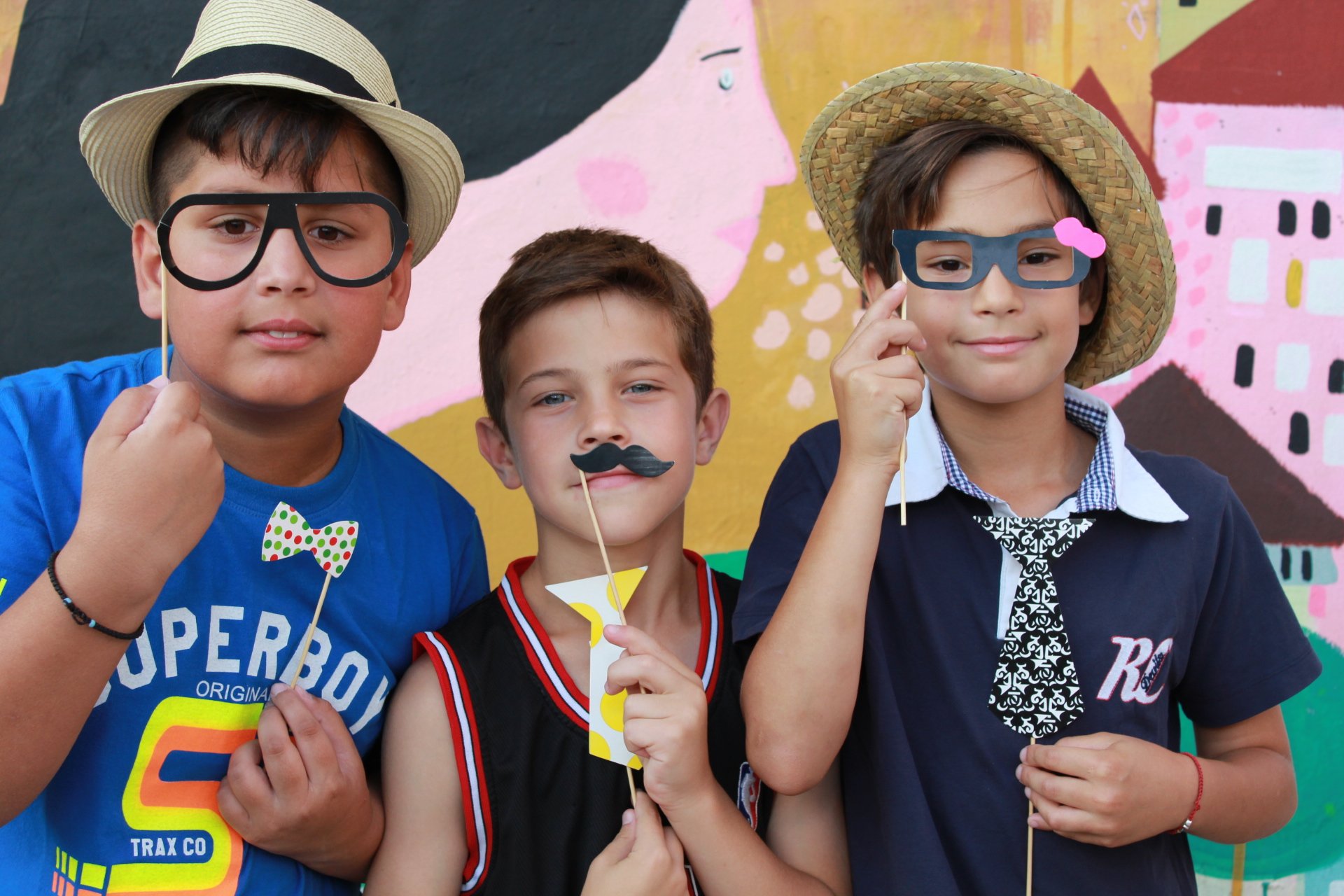 Three boys wearing hats and holding paper props like glasses, mustache, bow tie, and necktie in front of a colorful mural.